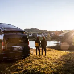 Un camping-car Westfalia avec panneaux solaires, prêt pour l'aventure près de Vulcania. Deux personnes contemplent un lac paisible au coucher du soleil.