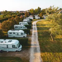 Vue aérienne d'un air camping car park, avec plusieurs camping-cars blancs alignés le long d'un chemin de terre, près d'un lac et d'une petite ville.