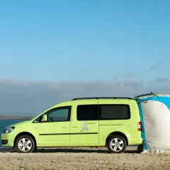 Un caddy tramper vert vif est garé sur une plage, avec une tente bleue et blanche attachée à l'arrière.