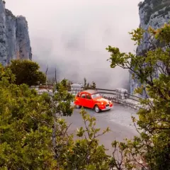 Une petite voiture rouge se gare sur la route des Gorges du Verdon, offrant une vue spectaculaire sur les falaises et la brume.