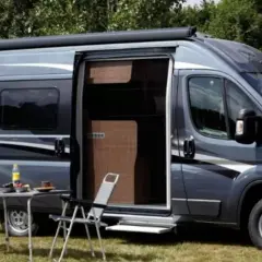 Un camping-car gris avec une moustiquaire plissée de porte ouverte. Une table et des chaises sont installées dehors.