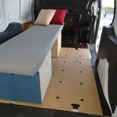 Aménagement intérieur d'un petit fourgon : banquette grise avec coussins, caisson de rangement bleu et blanc, plancher en bois. Prêt pour l'aventure !