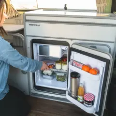 Une femme range des aliments dans un frigo pour van. Il contient des œufs, des légumes, des fruits et des boissons.