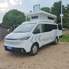 Ce camping-car 9m blanc, avec "Happy Life" écrit dessus, est prêt pour l'aventure. Il est garé sur un chemin de gravier, à côté d'une structure en bois.