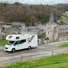 Un camping-car blanc roule dans un village pittoresque de Belgique, avec une église en pierre et des maisons anciennes.