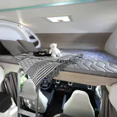 Lit de pavillon de camping-car avec oreillers, couverture et ours en peluche. Vue sur le poste de conduite.