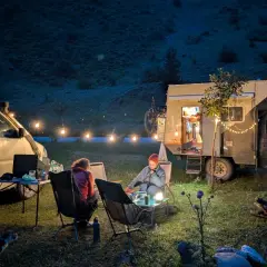 Soirée paisible autour d'un feu de camp, deux personnes assises près d'un **van aménagé électrique** illuminé par des guirlandes lumineuses.