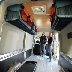 Aménagement fourgon par un professionnel : deux personnes admirent l'intérieur aménagé avec des rangements suspendus "Adventure Wagon" et un sol à rayures bleues.