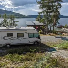 Un camping-car blanc est garé près d'un lac bordé de pins. Une petite cabane et un bateau sont visibles sur l'eau.