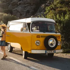A woman with a straw hat and camera leans against a yellow van, ready for an adventure in Majorque en van.