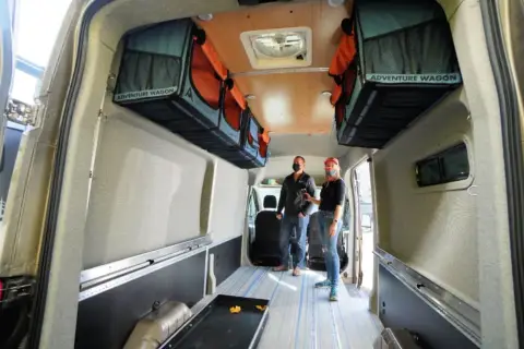 Aménagement fourgon par un professionnel : deux personnes admirent l'intérieur aménagé avec des rangements suspendus "Adventure Wagon" et un sol à rayures bleues.