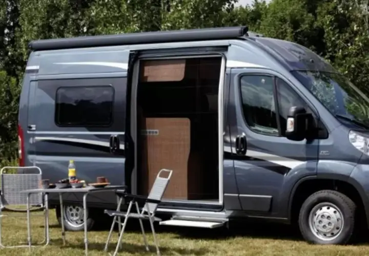 Un camping-car gris avec une moustiquaire plissée de porte ouverte. Une table et des chaises sont installées dehors.