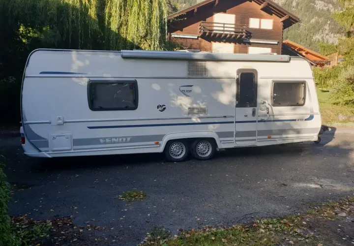 Caravane Fendt blanche garée devant une maison en bois, avec des arbres en arrière-plan.