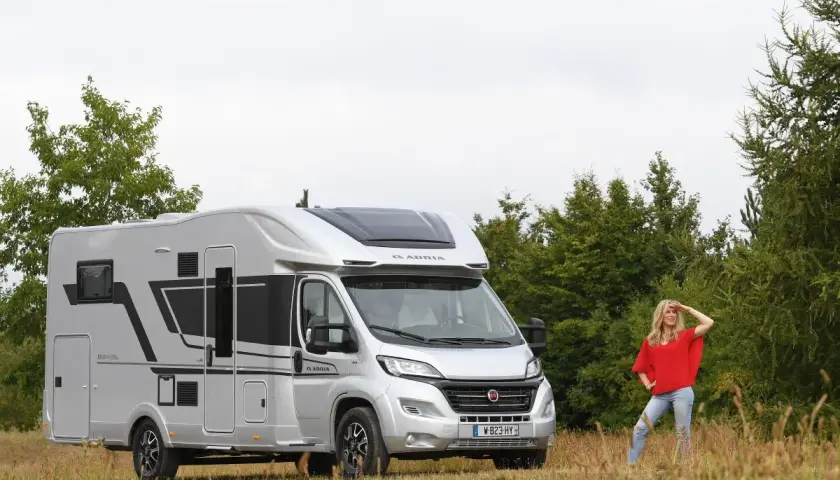 Une femme pose devant un camping-car Adria Matrix 670 SC argenté, prêt pour l'aventure.