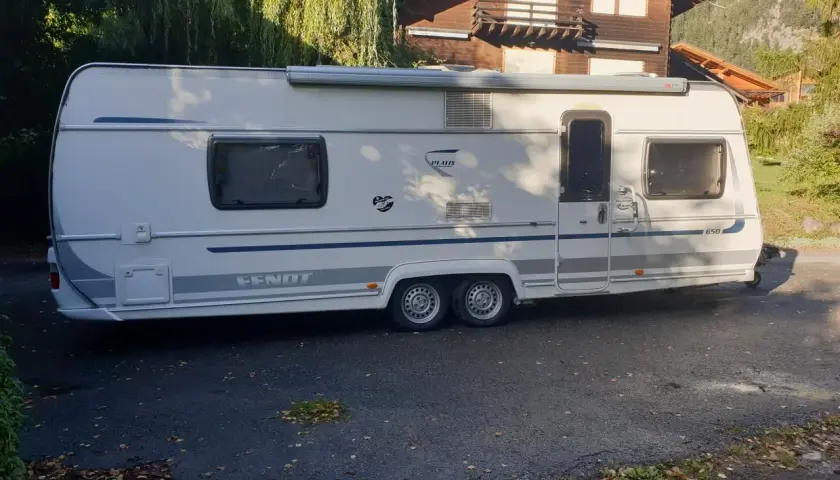 Caravane Fendt blanche garée devant une maison en bois, avec des arbres en arrière-plan.