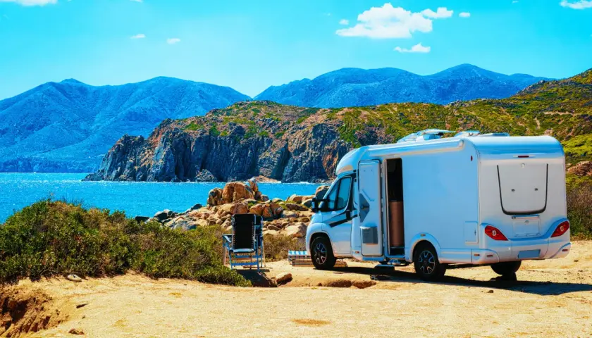 Un camping-car blanc est garé sur un terrain sablonneux, face à une mer bleue et des montagnes. L'application spot camping car idéale pour ce paysage.