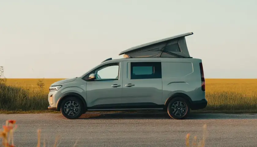 Un Dacia Sandman gris, prêt pour l'aventure, avec son toit relevable ouvert, stationné au bord d'un champ doré au coucher du soleil.