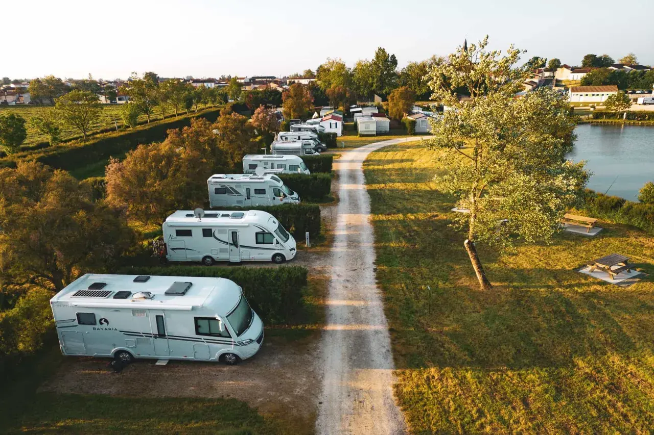 Vue aérienne d'un air camping car park, avec plusieurs camping-cars blancs alignés le long d'un chemin de terre, près d'un lac et d'une petite ville.