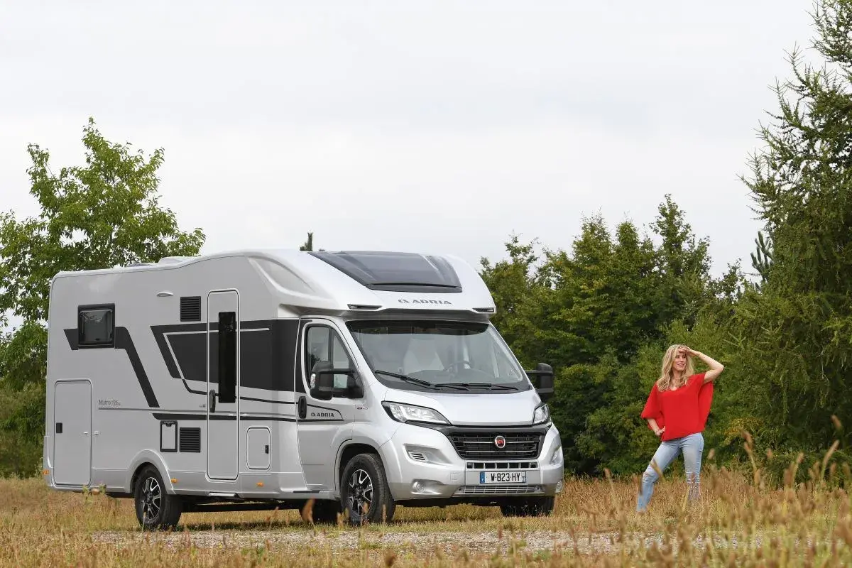 Une femme pose devant un camping-car Adria Matrix 670 SC argenté, prêt pour l'aventure.
