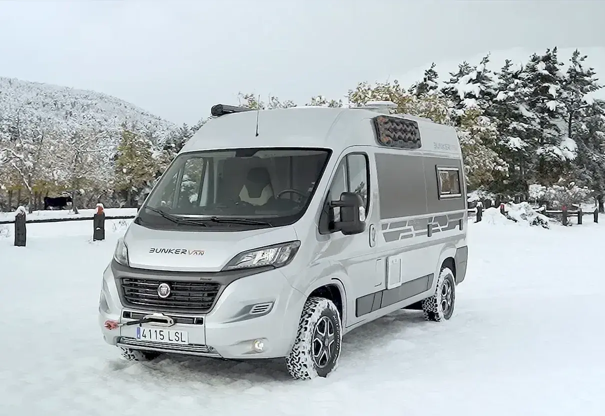 Camping-car 4x4 argenté garé dans la neige, avec des arbres enneigés en arrière-plan.