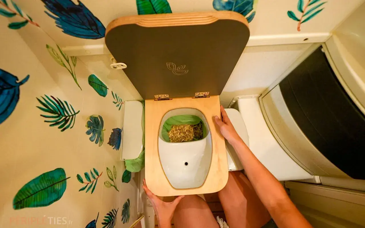 Vue plongeante d'une toilette sèche avec couvercle ouvert, remplie de copeaux. Les murs sont décorés de motifs de feuilles tropicales. Les toilettes sèches inconvénients peuvent inclure la gestion des matières.