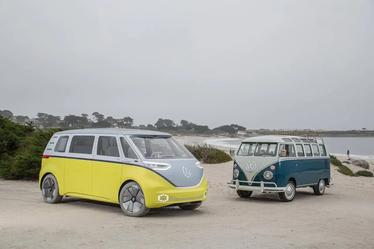 Deux combi VW, un moderne jaune et gris, l'autre vintage bleu et blanc, sur une plage brumeuse.