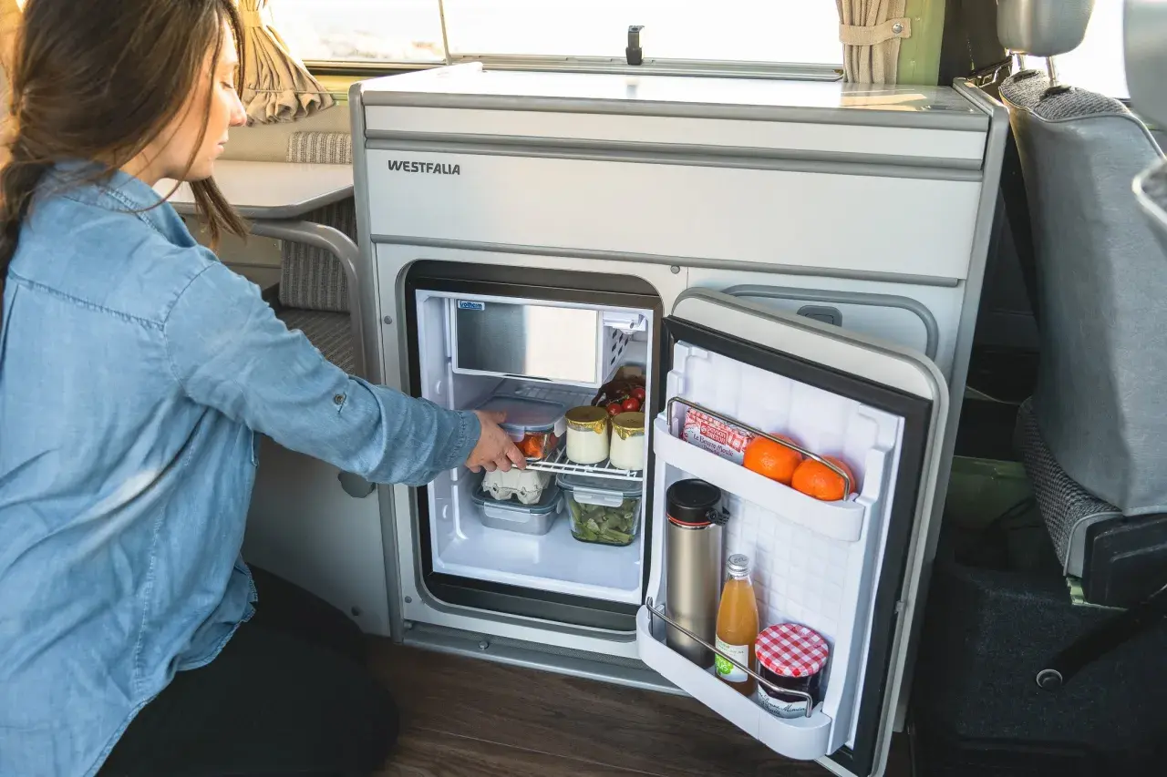 Une femme range des aliments dans un frigo pour van. Il contient des œufs, des légumes, des fruits et des boissons.