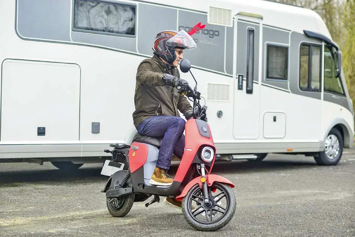 Un homme casqué conduit un scooter rouge devant un camping-car. Ce scooter est parfait pour explorer les environs lors de vos voyages en camping-car.
