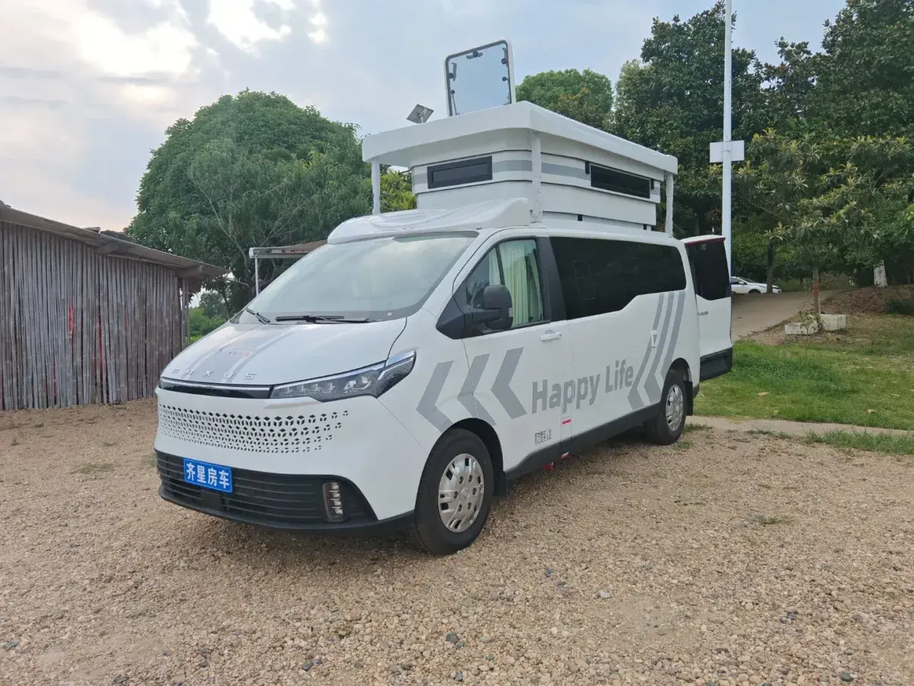Ce camping-car 9m blanc, avec "Happy Life" écrit dessus, est prêt pour l'aventure. Il est garé sur un chemin de gravier, à côté d'une structure en bois.