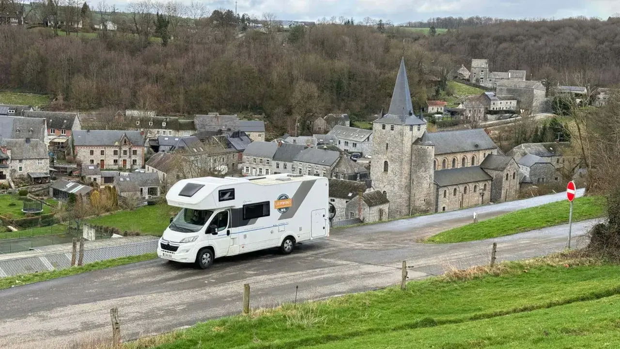 Un camping-car blanc roule dans un village pittoresque de Belgique, avec une église en pierre et des maisons anciennes.