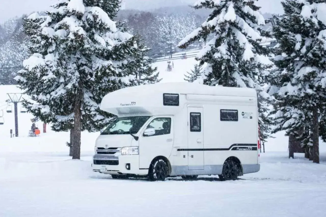 Camping car japonais blanc garé dans la neige, entouré de sapins enneigés. Des remontées mécaniques sont visibles en arrière-plan.