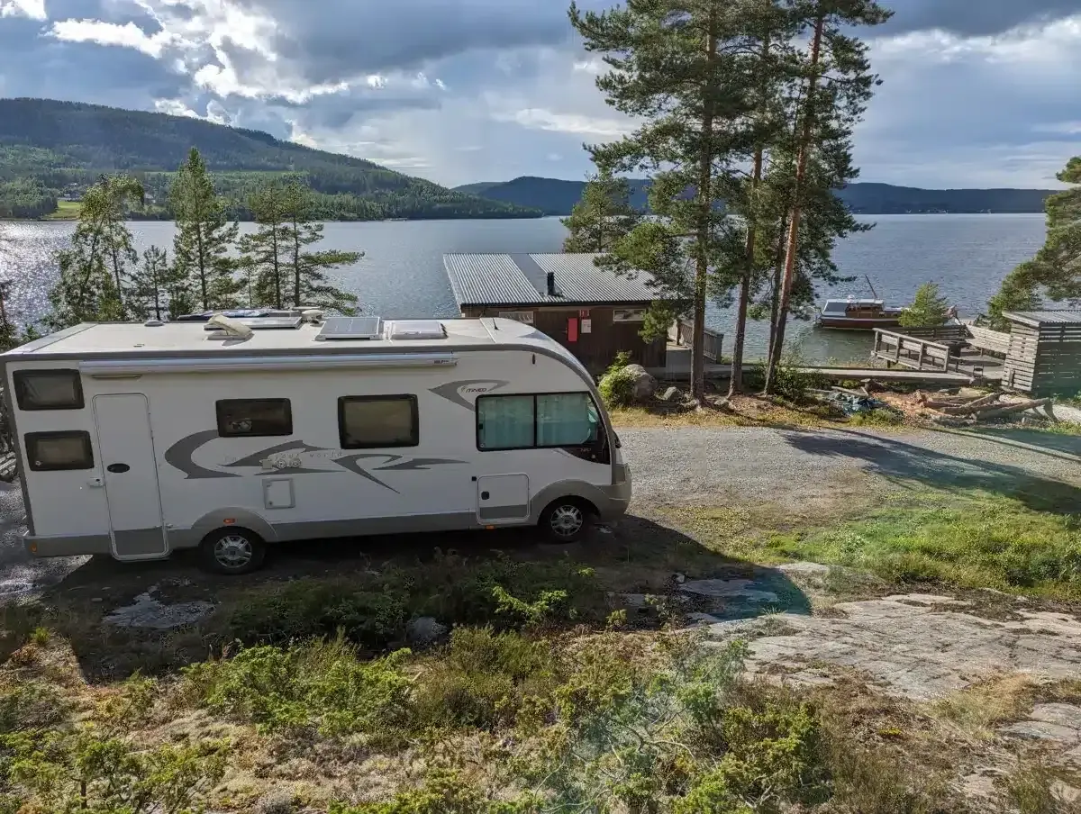 Un camping-car blanc est garé près d'un lac bordé de pins. Une petite cabane et un bateau sont visibles sur l'eau.