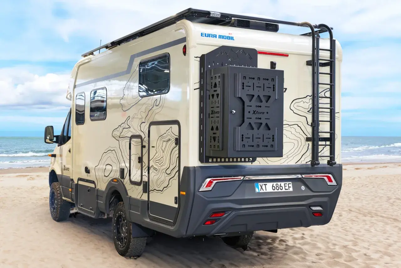 Camping-car Eura Mobil Xtura sur une plage de sable, avec une échelle arrière et un porte-roue noir.