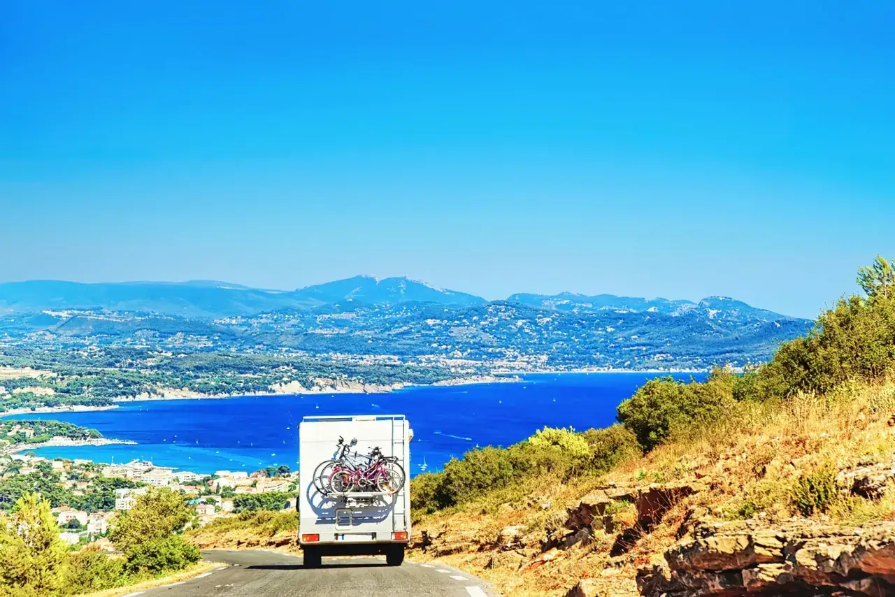 Un camping-car avec v&eacute;los &agrave; l'arri&egrave;re descend une route sinueuse vers une baie bleue. Parfait pour une id&eacute;e sortie week-end camping-car.