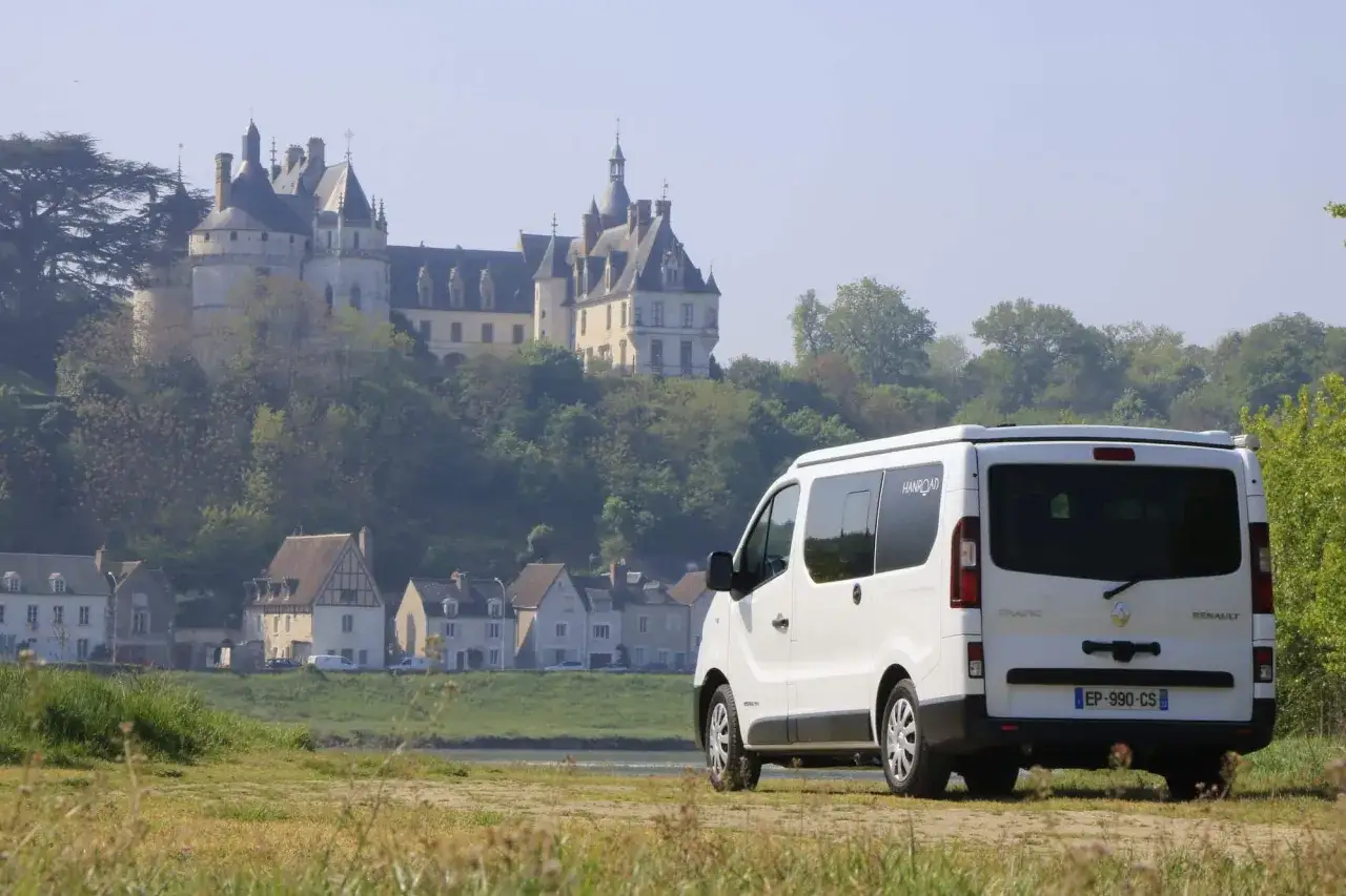 Camping-car blanc gar&eacute; devant un village pittoresque et des ch&acirc;teaux allemands majestueux perch&eacute;s sur une colline verdoyante.