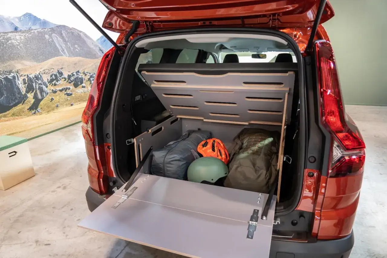 Coffre ouvert d'un camping car Dacia, aménagé avec des sacs de couchage, casques et une table rabattable. Prêt pour l'aventure !