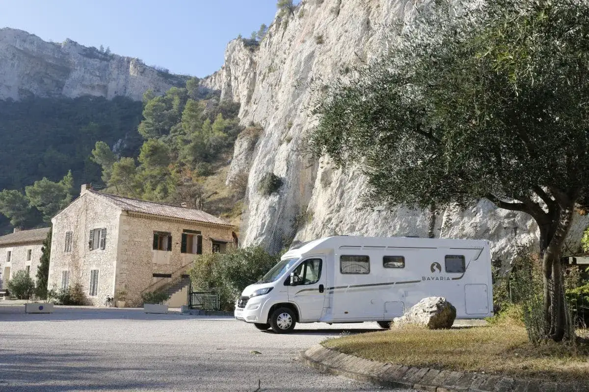 Un camping-car Bavaria blanc est gar&eacute; devant une maison en pierre, sous une falaise imposante, dans un d&eacute;cor typique de la Provence.