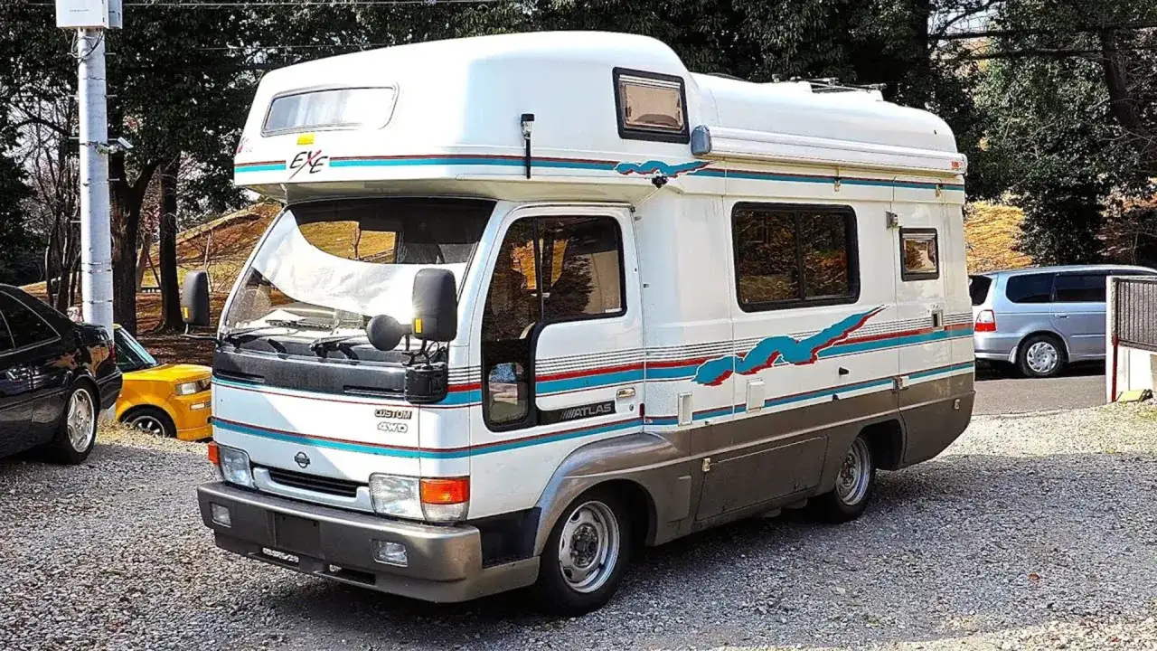 Un camping-car japonais blanc et gris, avec des bandes bleues et rouges, prêt pour l'aventure.