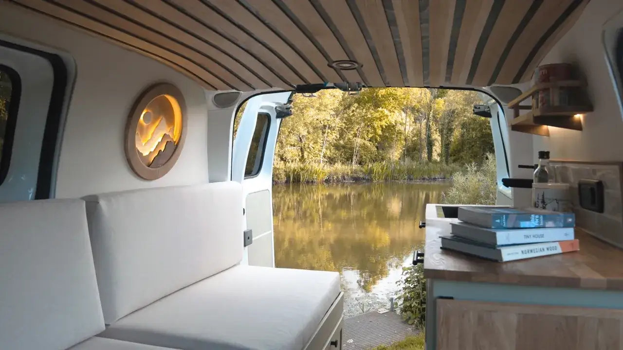 Int&eacute;rieur d'un camping car &eacute;lectrique avec vue sur un lac. Canap&eacute; blanc, plafond en bois, et une d&eacute;coration murale de montagne.