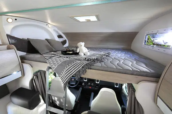 Lit de pavillon de camping-car avec oreillers, couverture et ours en peluche. Vue sur le poste de conduite.
